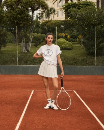A woman stands on a red tennis court wearing a white t-shirt, pleated tennis skirt, white tennis shoes, and holding a tennis racket.