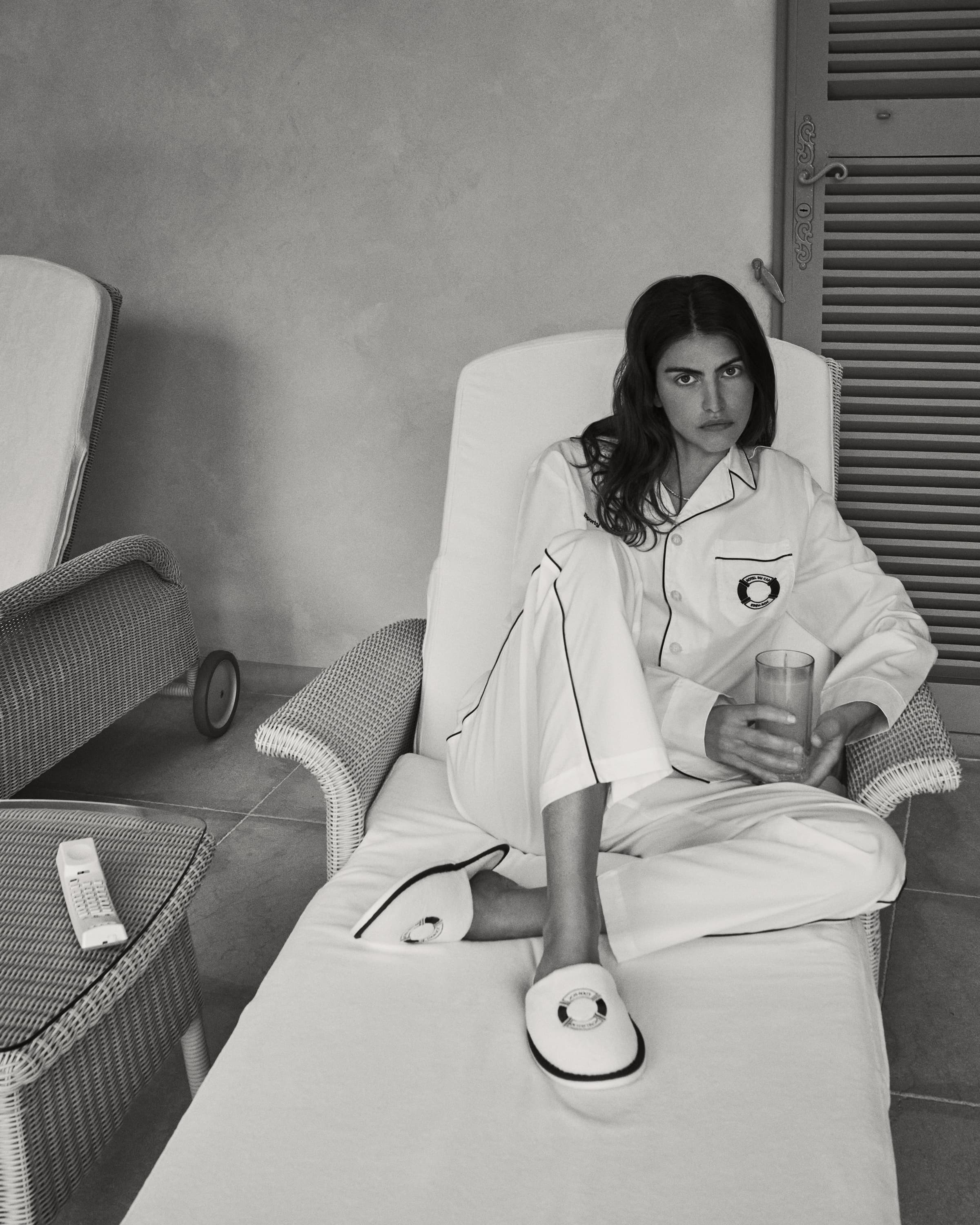 Monochrome shot of a woman in white pajamas sitting on a lounge chair, holding a glass, wearing slippers in an indoor setting.