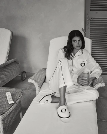 Monochrome shot of a woman in white pajamas sitting on a lounge chair, holding a glass, wearing slippers in an indoor setting.