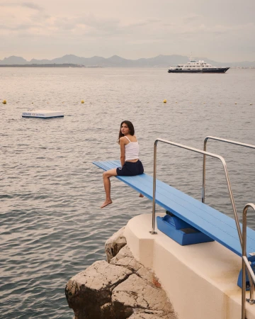 A young woman sits on a diving board overlooking the ocean with a yacht in the distance on a cloudy day, capturing a serene moment.