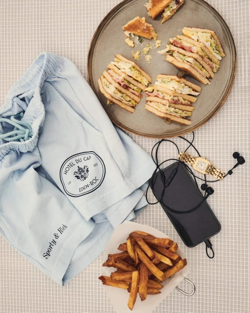 Overhead shot of light blue shorts, layered sandwiches, french fries, a black smartphone, wired earbuds, and a gold watch on a tablecloth.