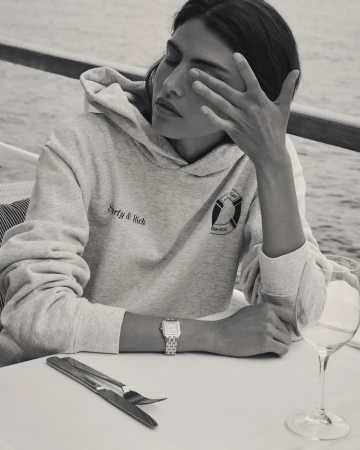 A woman in a gray hoodie at a table with silverware and a wine glass, looking off in the distance with hand near her face.