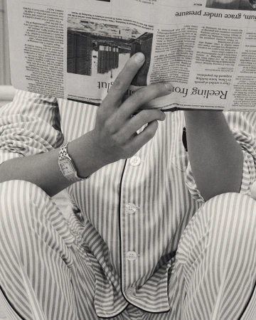 Person in striped pajamas sits cross-legged while reading a newspaper that covers their face, monochrome photo.