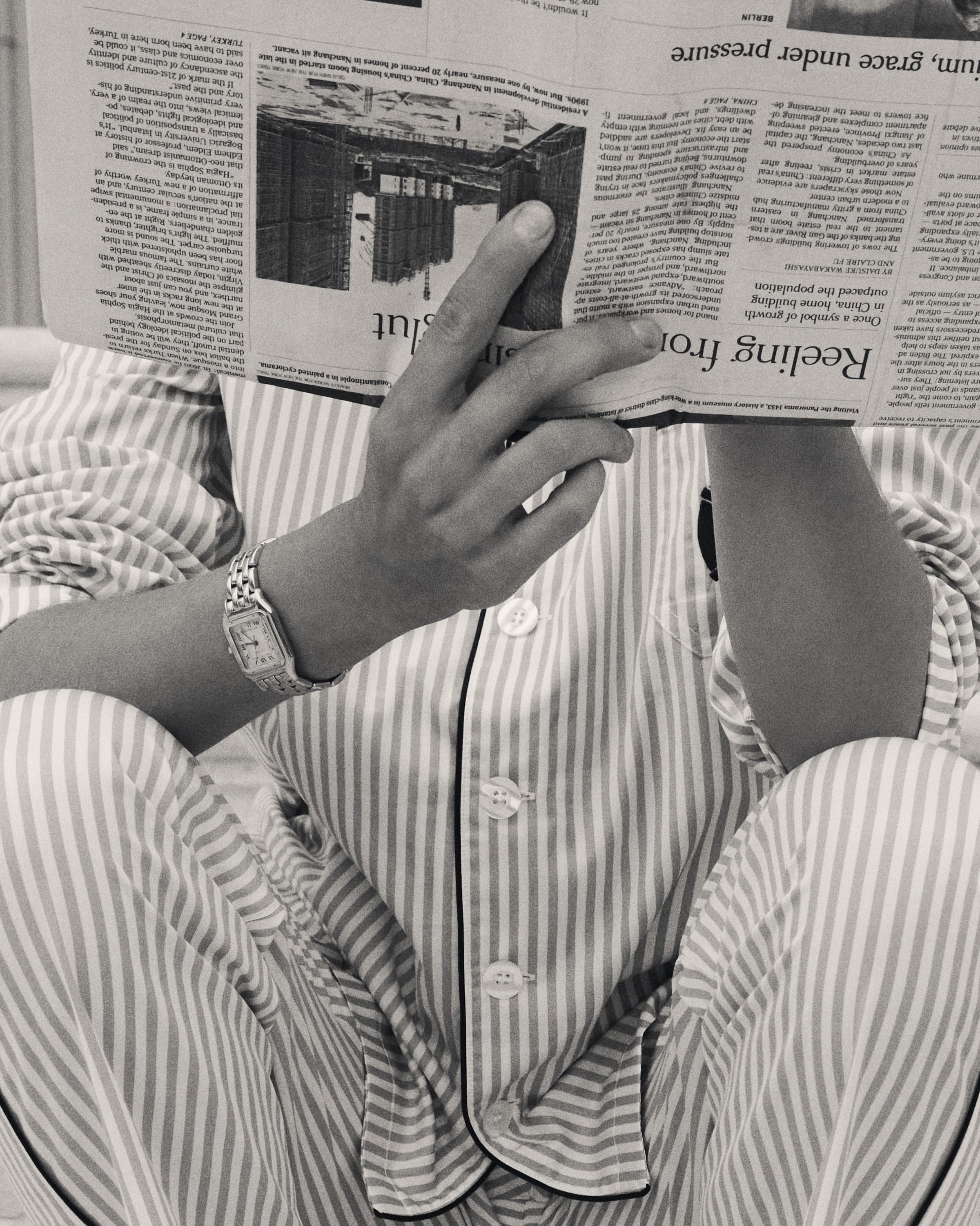 Person wearing a striped pajama set and silver wristwatch holding an open newspaper in front of their face in black and white.