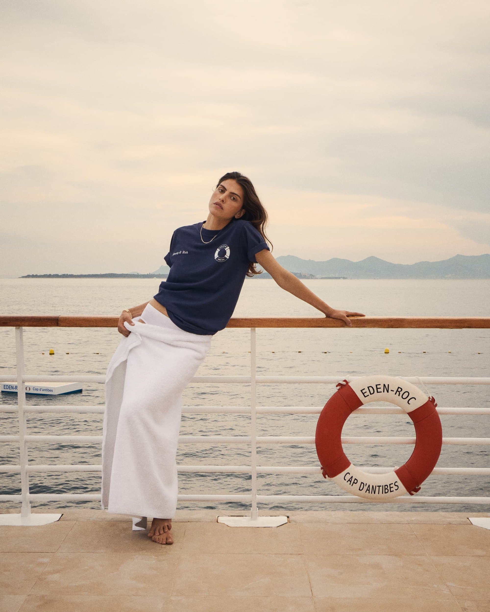 Model leaning on a seaside deck railing wearing a navy blue t-shirt and white towel wrap near an Eden-Roc lifebuoy.