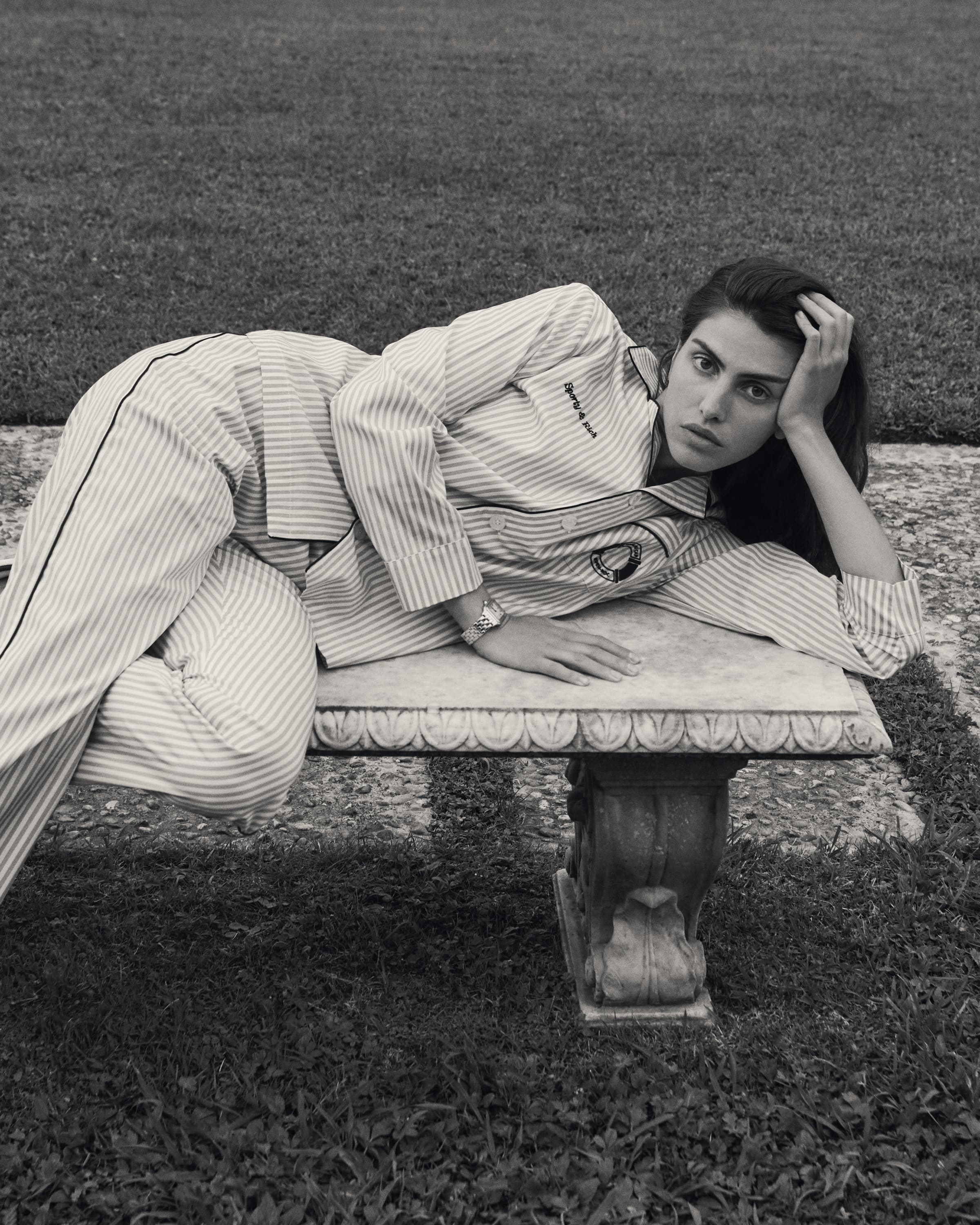 Woman in a striped pajama set reclining on an ornate stone garden bench outdoors in black and white.