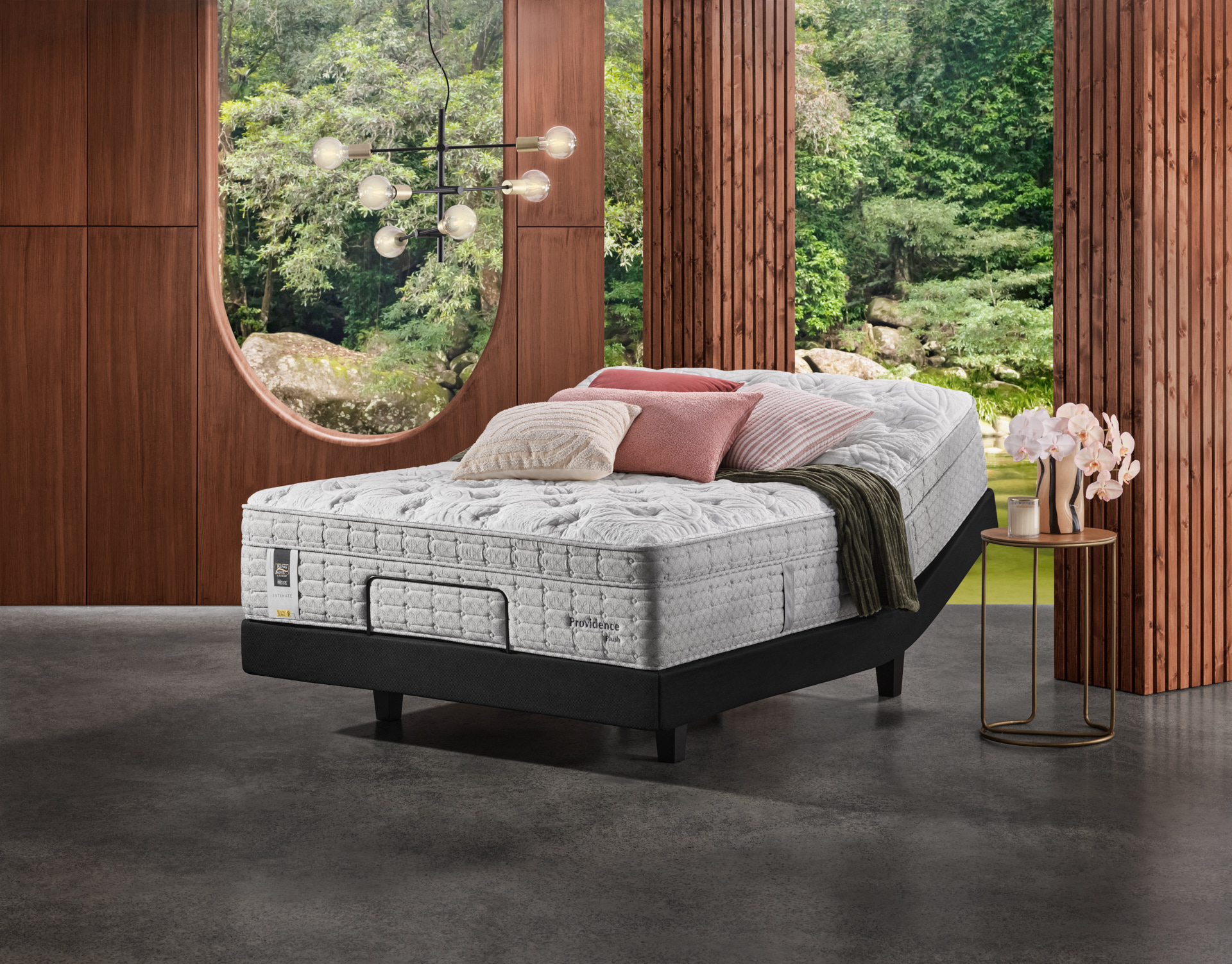 Elevated adjustable bed with pillows on a black platform frame indoors, bedside table with flowers, and a forest visible through a window.