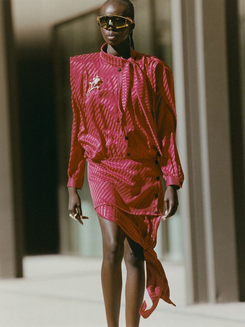 A woman in a pink striped dress and gold sunglasses confidently strides outdoors on a sunny day, showcasing a striking fashion statement.