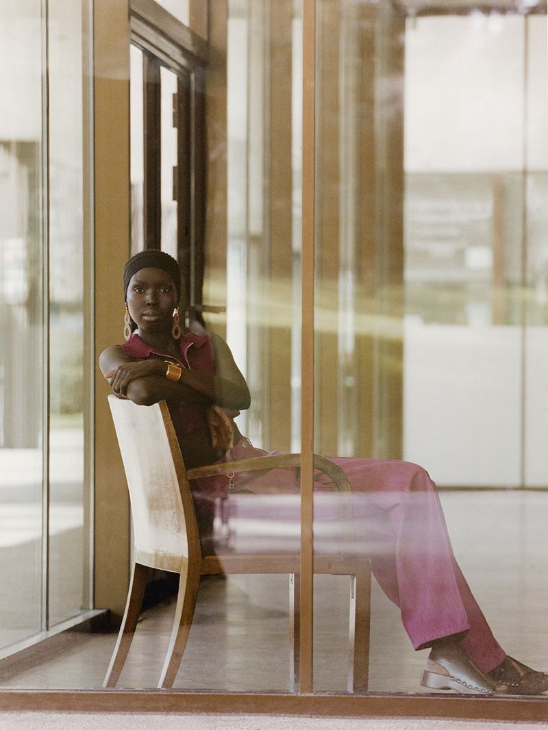 Portrait of a stylish African woman in a maroon outfit sitting in a chair with a modern architectural background, posing elegantly.