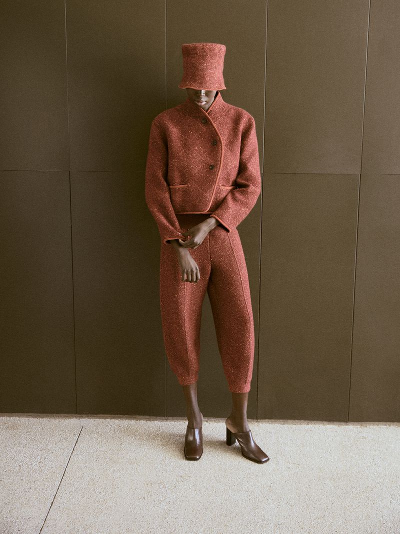 Fashion shot of a Black female model in a rust-colored cropped blazer, wide leg trousers, and bucket hat, posed against a neutral wall.