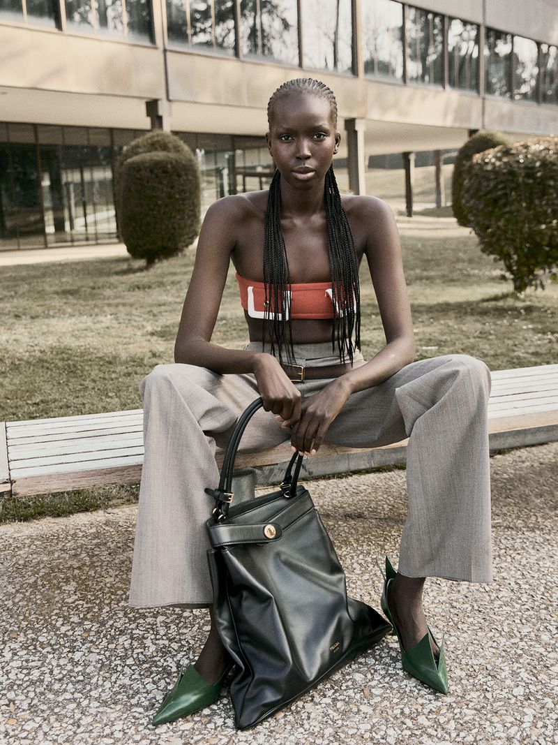 Fashion shot of a dark-skinned woman with braids, wearing an orange tube top and wide trousers, sitting on a bench holding a leather bag.