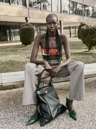 Fashion shot of a dark-skinned woman with braids, wearing an orange tube top and wide trousers, sitting on a bench holding a leather bag.