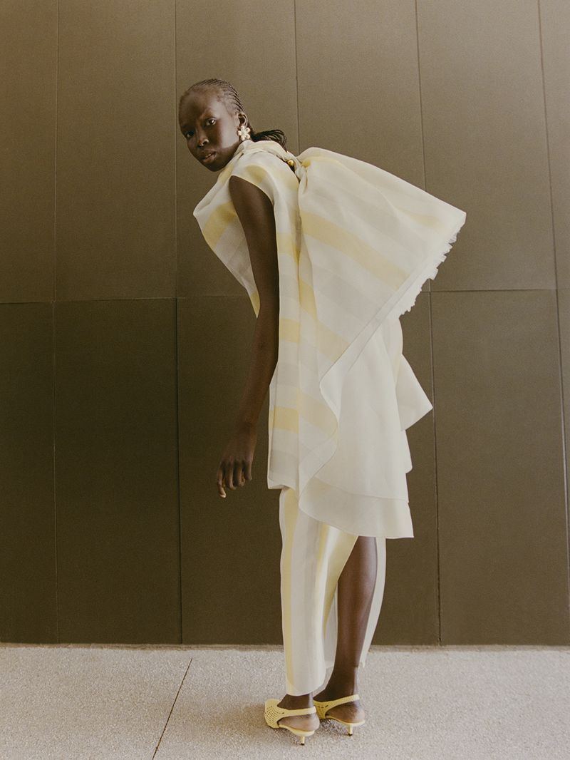 An elegant Black female model poses gracefully in a flowing white and yellow striped dress, complemented by yellow heels in a studio setting.