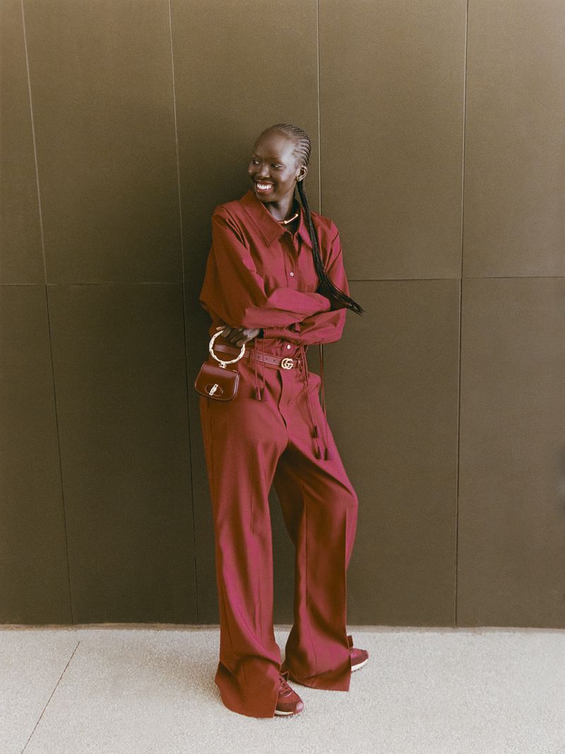 Portrait of a Black woman in a dark red pantsuit leaning against a textured wall, smiling, with a small purse at her hip.