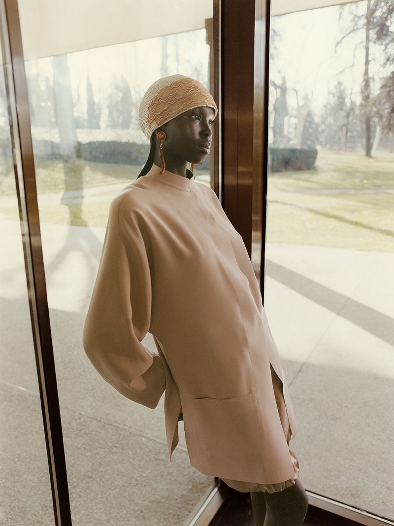 A dark-skinned model poses in a light peach-colored outfit and matching head covering near large glass windows with a blurred outdoor view.