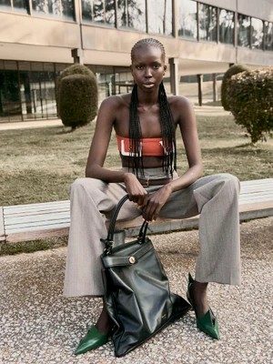 Model Ajok Madel seated on a wooden bench wearing grey trousers, a bandeau top, and holding a large black handbag.