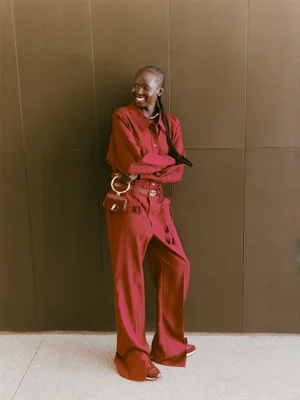 Model Ajok Madel wearing a burgundy matching outfit holding a small handbag against a panelled wall