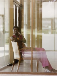 Model Ajok Madel seated on a wooden chair behind glass panels wearing a pink outfit for Harper's Bazaar Espana