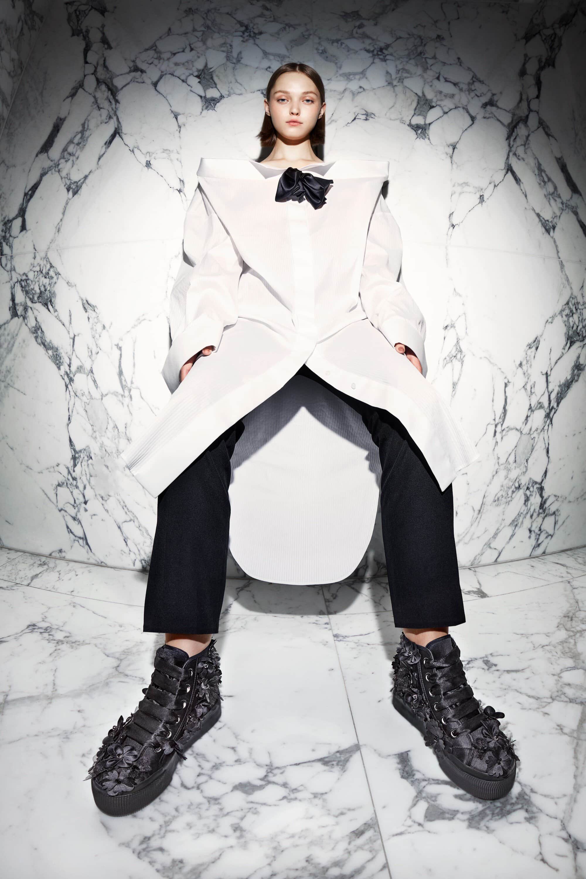 Young woman poses in a dramatic wide shot wearing black pants, platform sneakers, and a white, oversized shirt with a black bow.