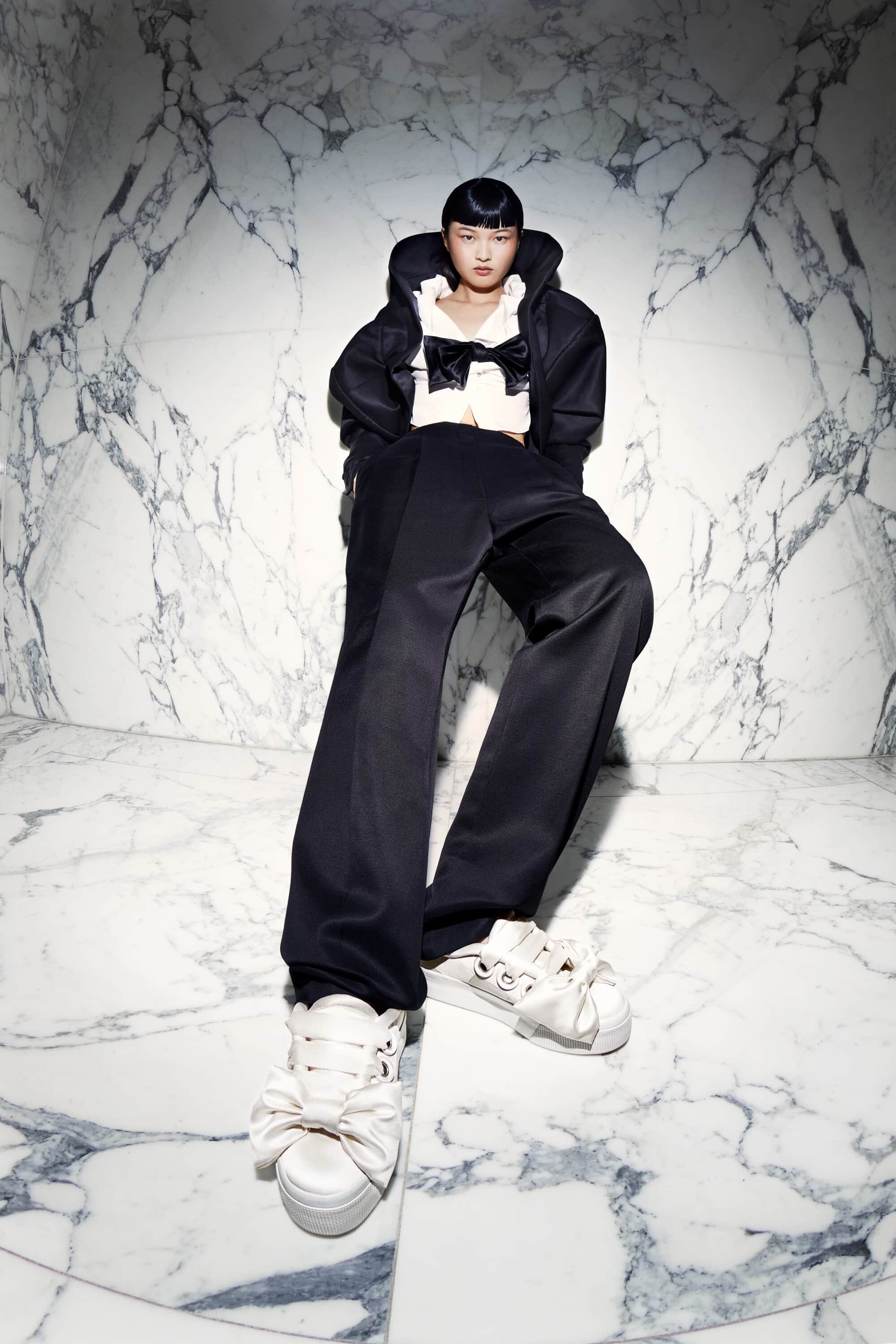 Woman in a black pantsuit with a white blouse and sneakers with bows poses against a marble wall in a studio setting.
