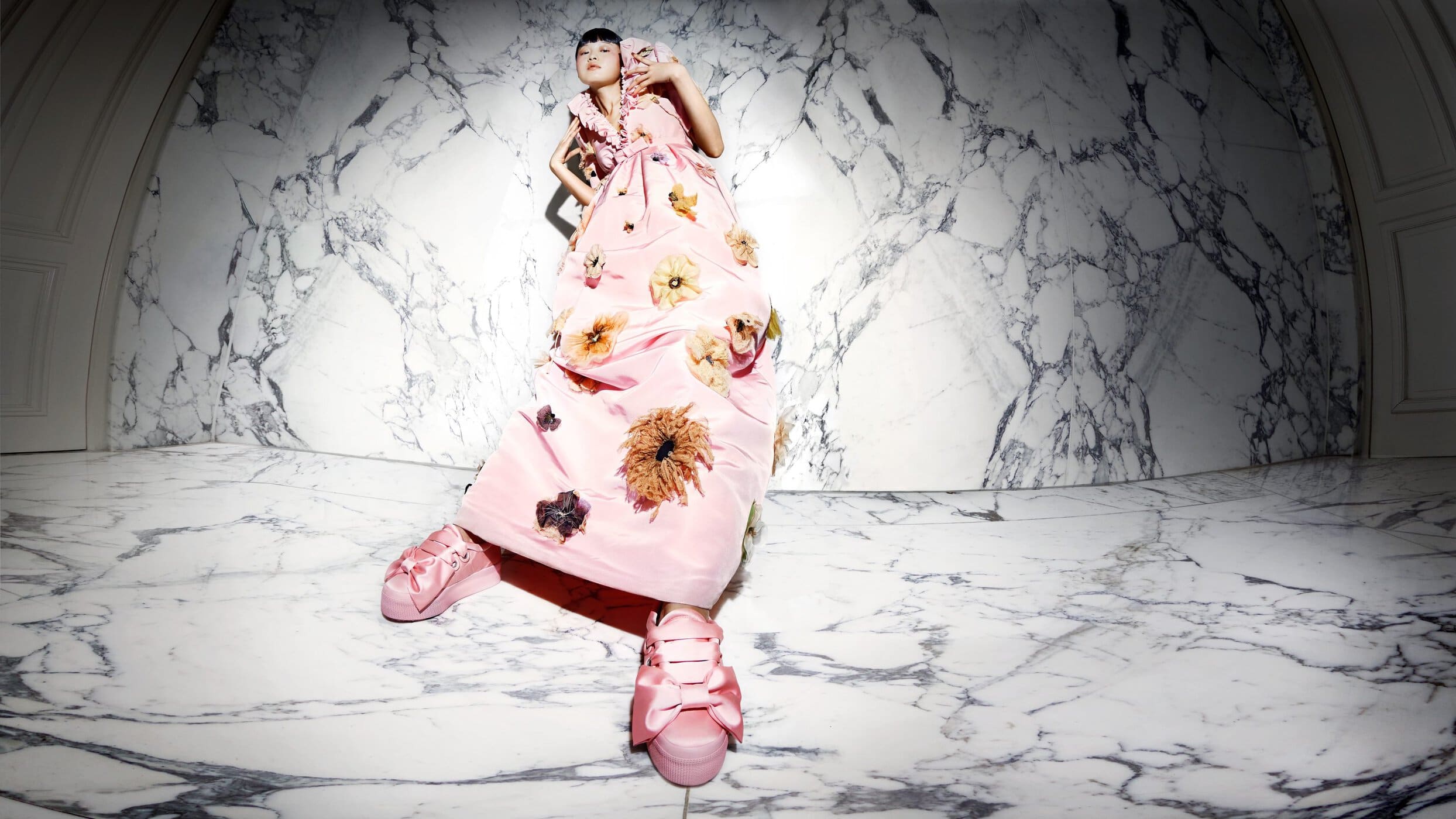 A fashionable Asian woman lying on a marble floor wears a pink floral dress and matching sneakers in an indoor studio setting.