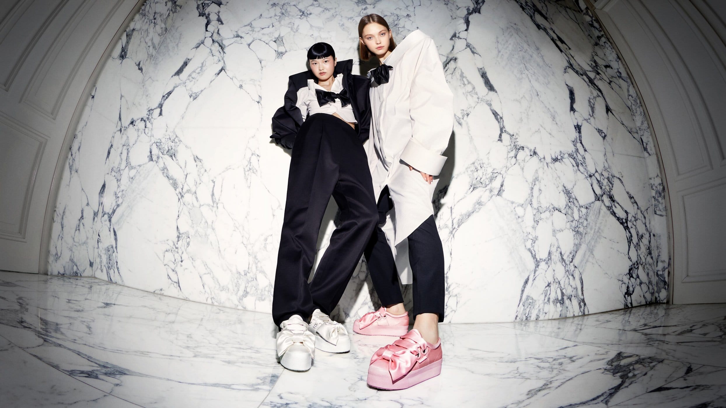 Two fashion models strike a pose wearing oversized bow-tie tops, black trousers, and platform sneakers against a marble backdrop.