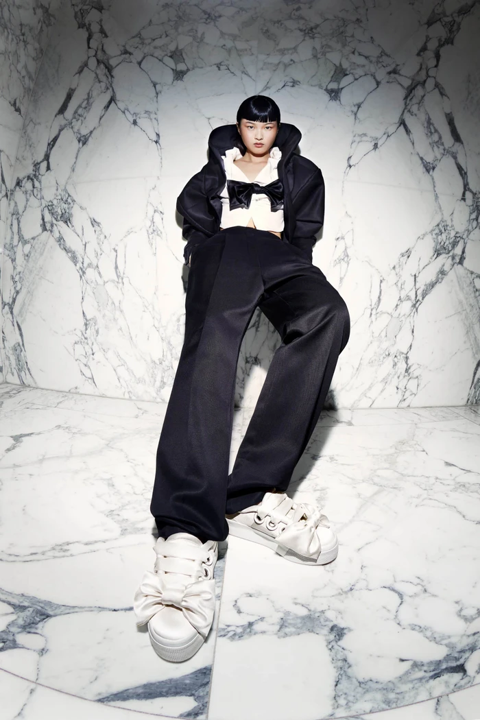 Model in black jacket and trousers with white sneakers featuring large bows against a white marble wall.