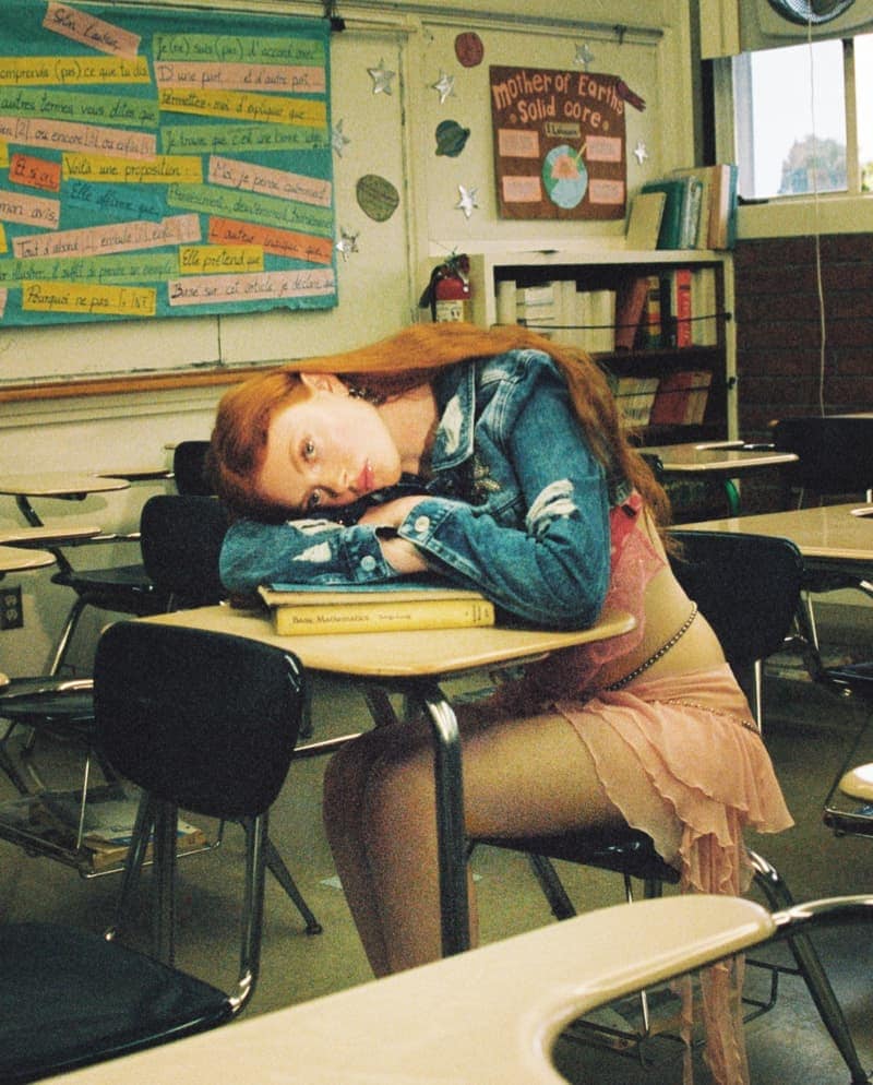 Red-haired woman in a denim jacket rests her head on books on a school desk in a classroom with desks and learning materials.