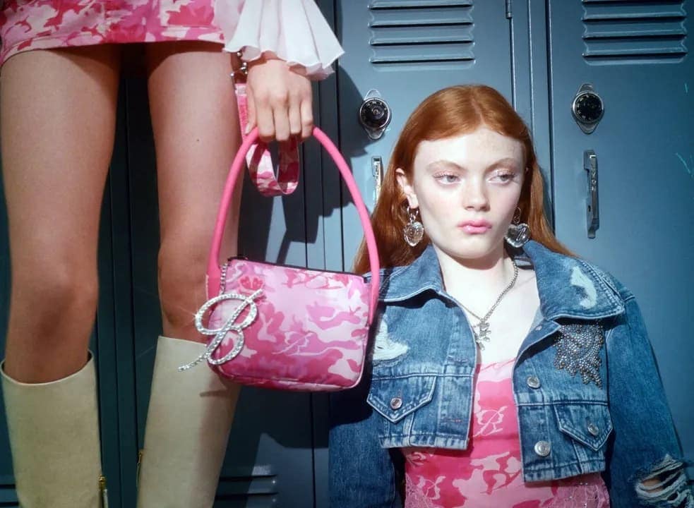 Two women in a high school hallway with lockers, one holding a pink purse with a 'B' charm, both wearing pink camo outfits.
