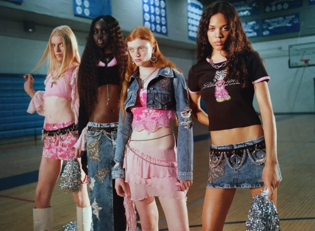 Four women in trendy Y2K outfits stand in a gymnasium, posing for a fashion shoot. The women wear crop tops, miniskirts, and platform boots.