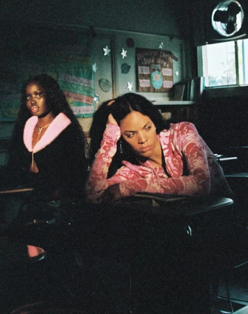 Two women in a classroom setting. One woman wears a pink floral top with her head in her hand and the other is wearing black