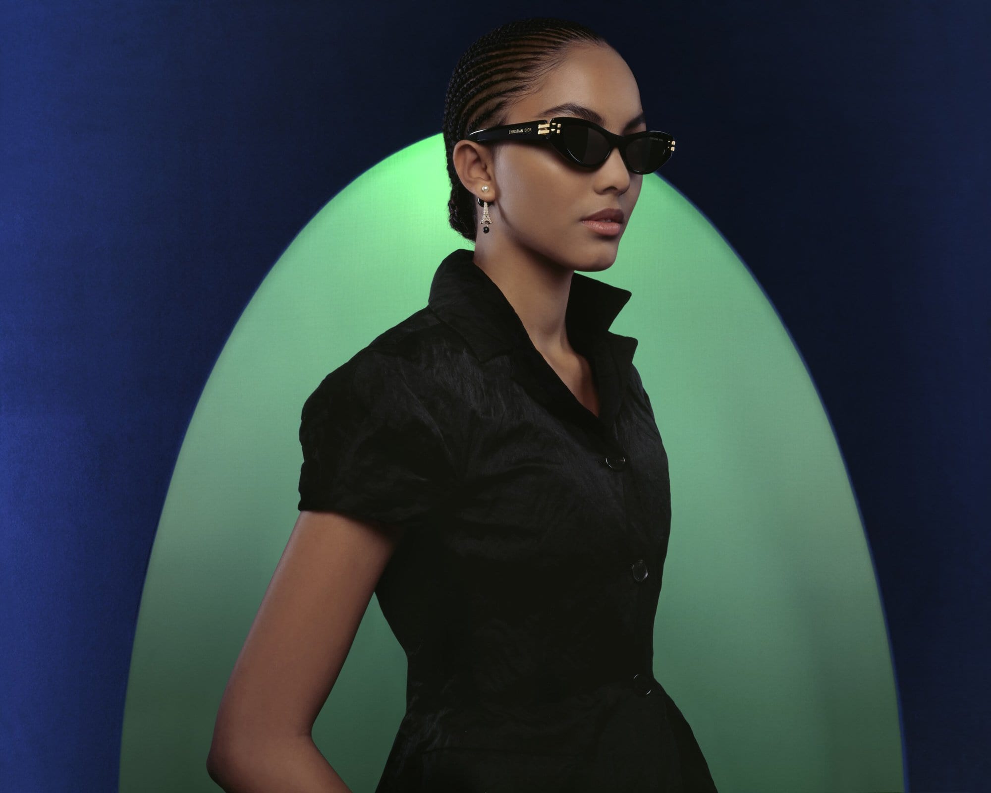 Model wearing dark sunglasses and black collared shirt against a blue and green arched studio backdrop for Dior Fall 2023 campaign.