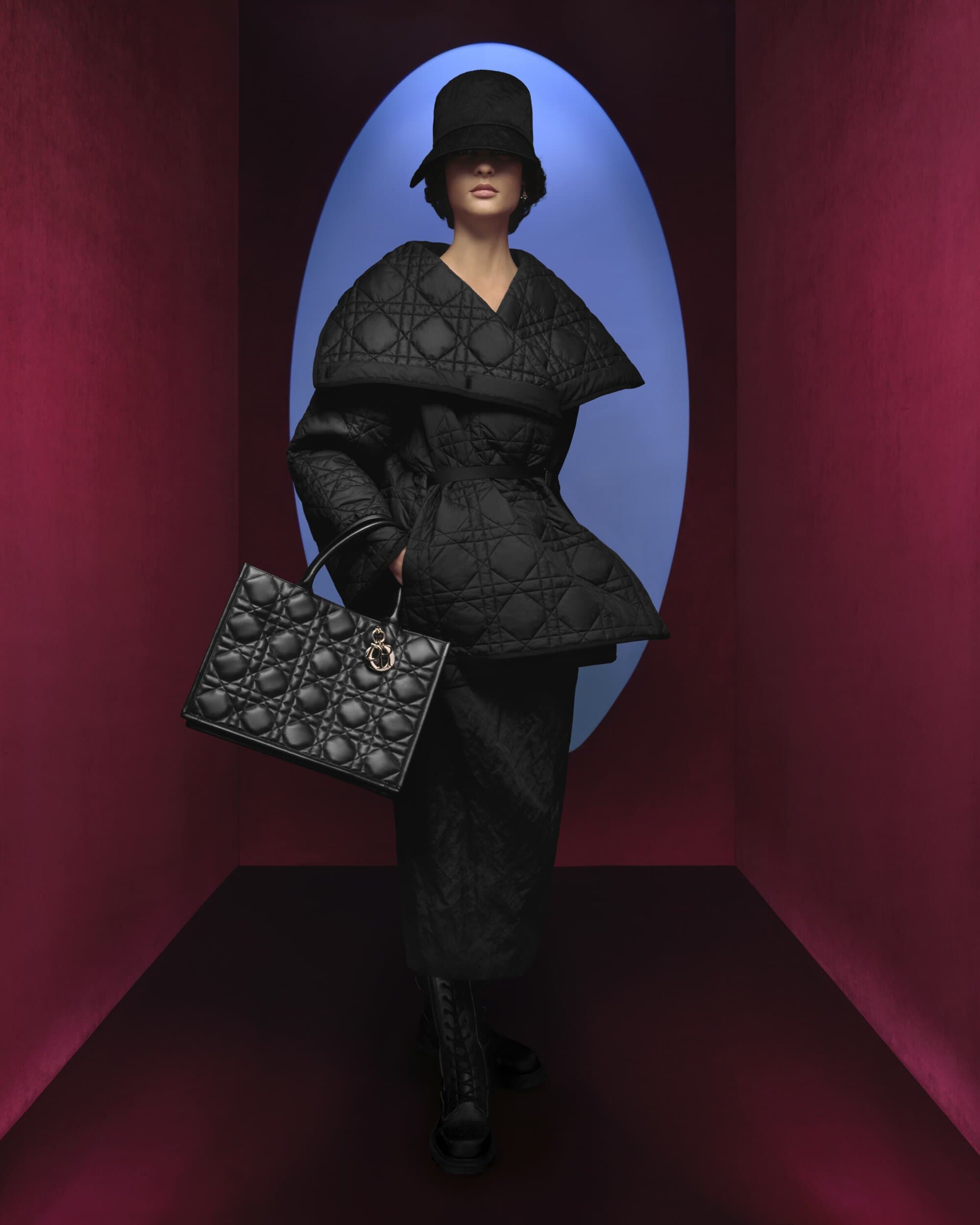 Elegant adult female fashion model in black designer coat, hat, skirt and handbag against a blue and burgundy background.