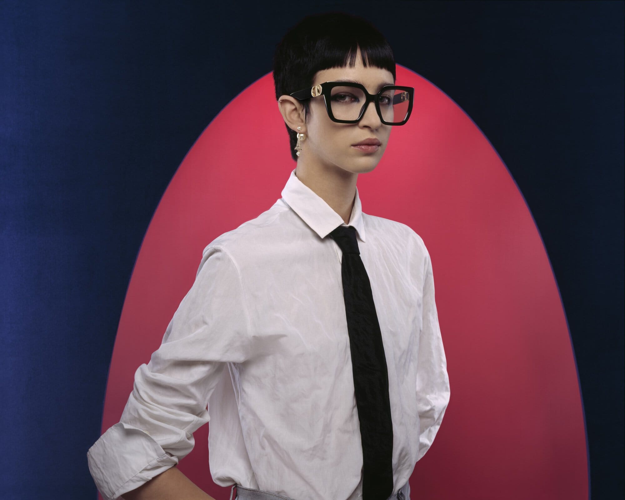 A young adult woman wearing glasses and a tie poses indoors against a duotone backdrop in a fashion-forward studio portrait.