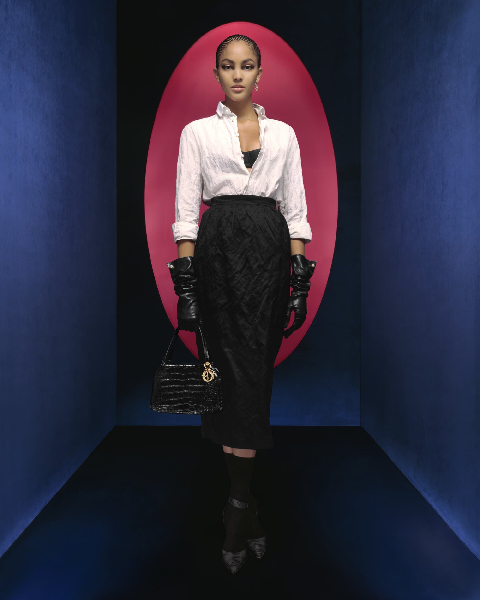 Fashionable woman in a white shirt, black skirt, and gloves poses with a handbag against a vibrant blue and pink studio backdrop.