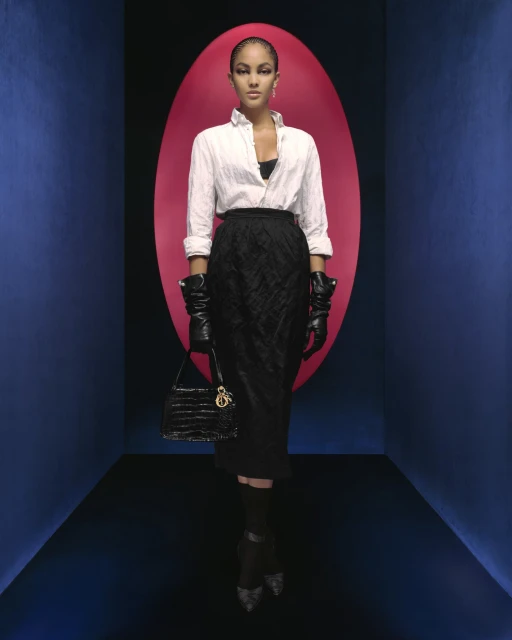 Fashionable woman in a white shirt, black skirt, and gloves poses with a handbag against a vibrant blue and pink studio backdrop.