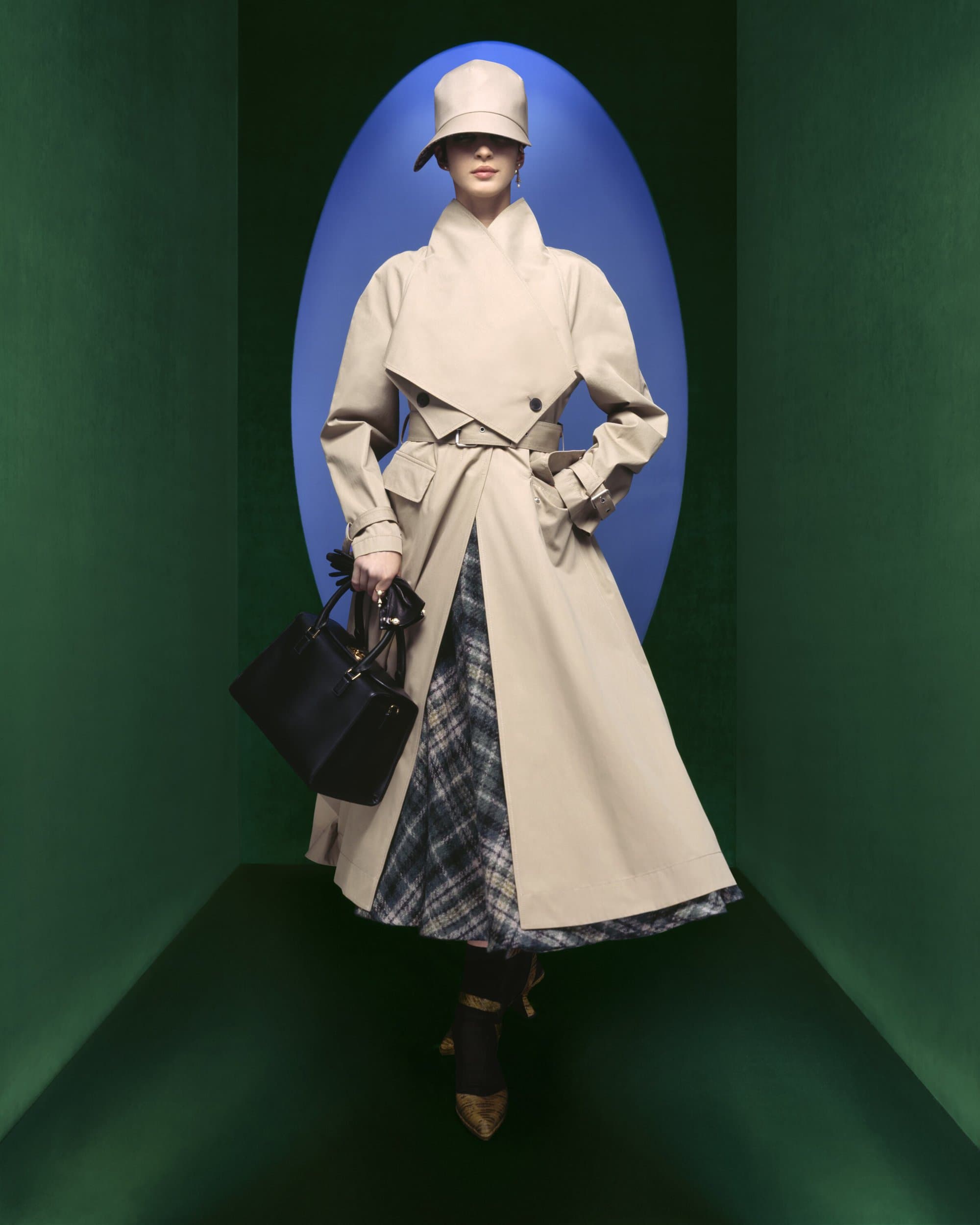 Model wearing a beige structured trench coat, matching cloche hat, and holding a black handbag in a green frame with a blue oval.