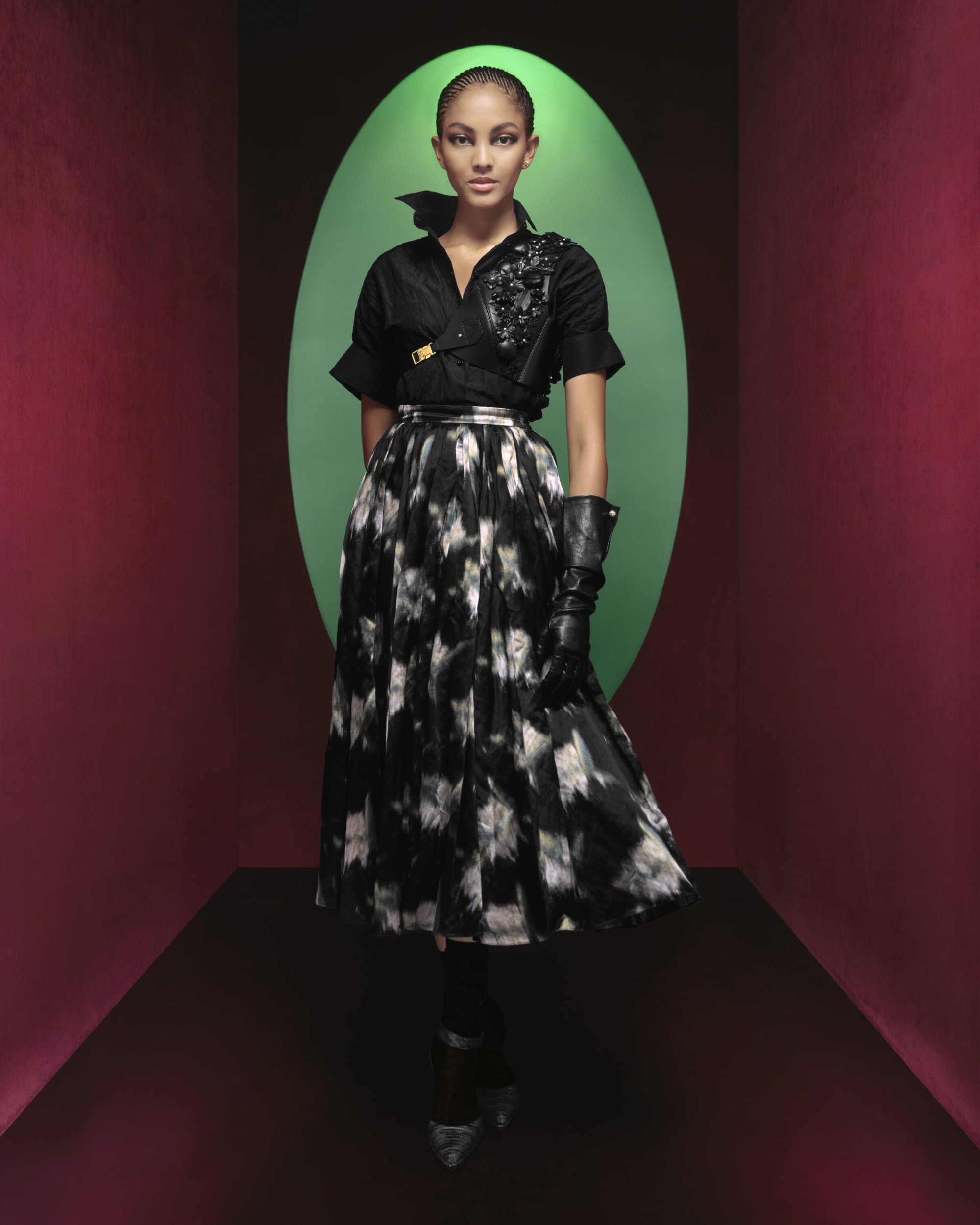 Fashion shot of a model in a black blouse, patterned skirt, and leather gloves posing in front of a green oval backdrop.