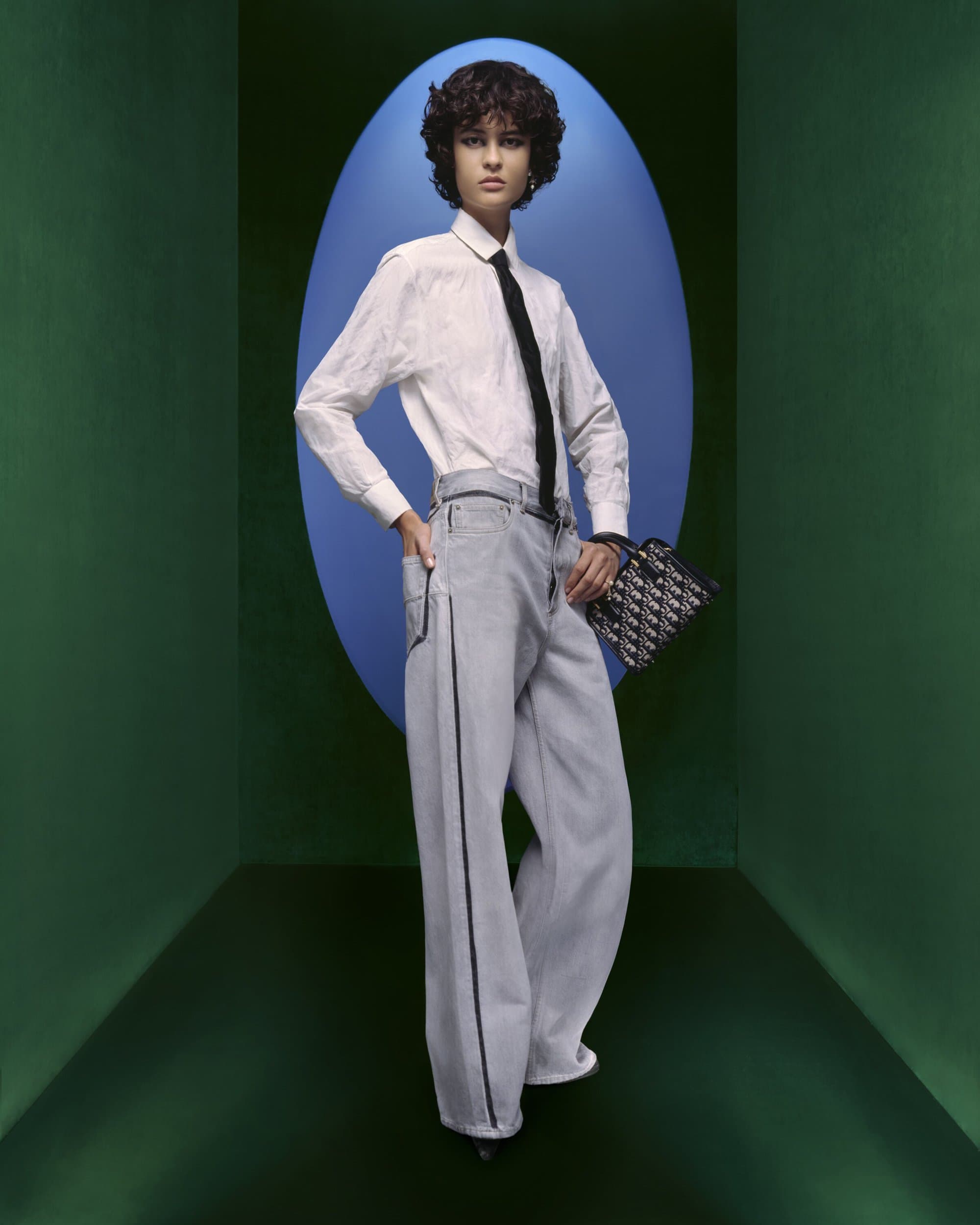 Fashion shot of a woman in a button-down shirt, wide-leg pants, and a tie holding a purse in a teal and blue geometric set.
