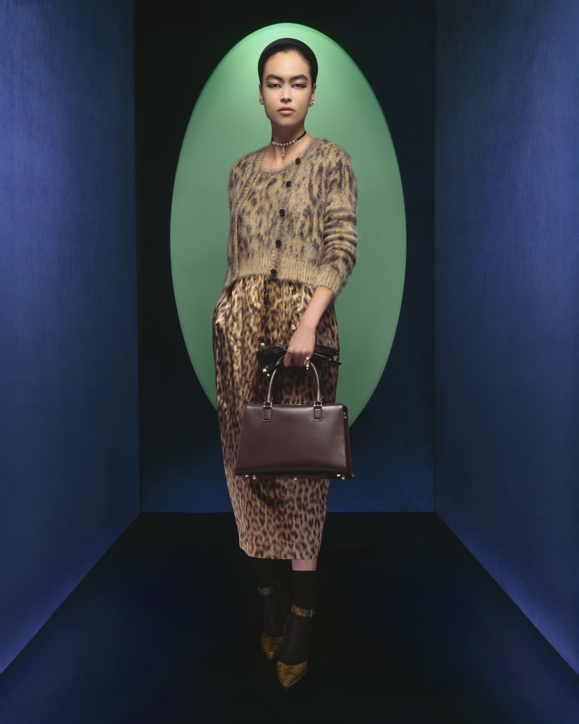 A model showcases a brown leather handbag paired with a leopard print skirt and cardigan in a studio setting with blue and green walls.