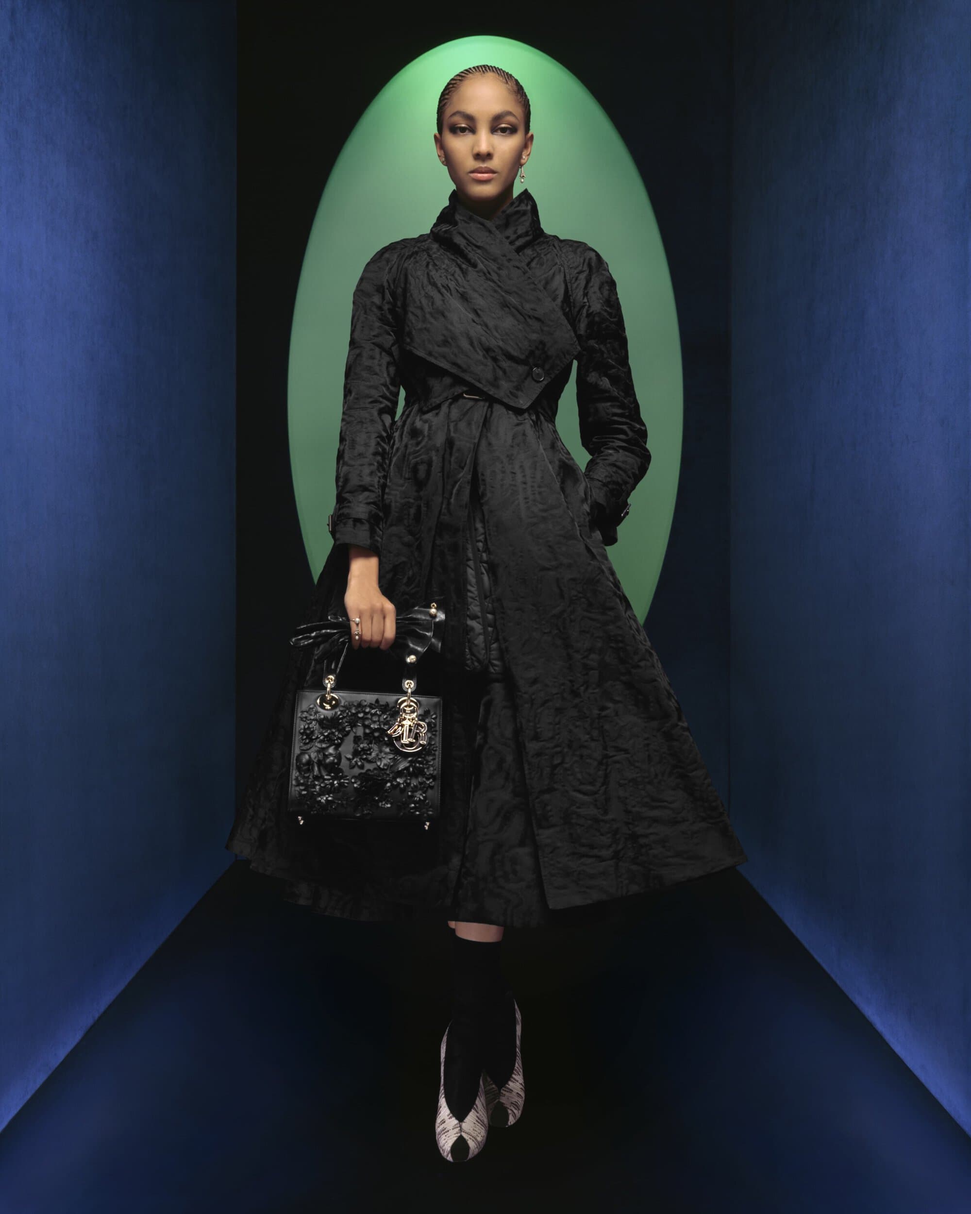 Model in black textured coat holding a dark handbag standing before a green oval archway inside a blue tunnel
