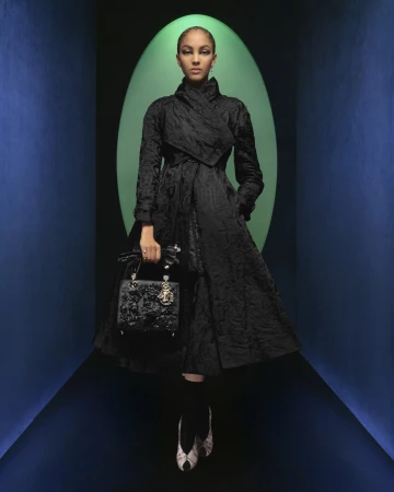Model in black textured coat holding a dark handbag standing before a green oval archway inside a blue tunnel