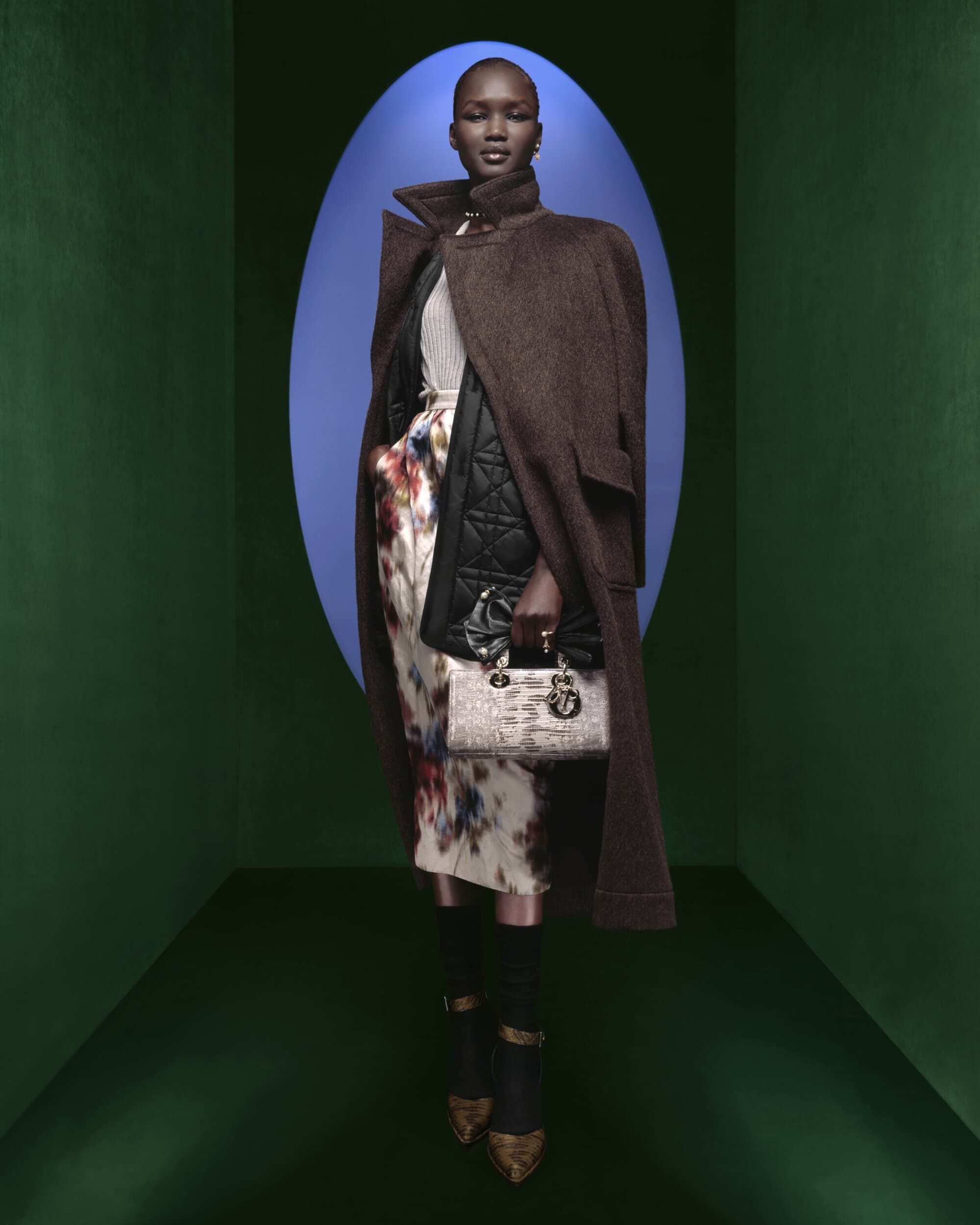 Full-length portrait of a Black female fashion model in a brown coat, floral dress, and black handbag, against a blue and green backdrop.
