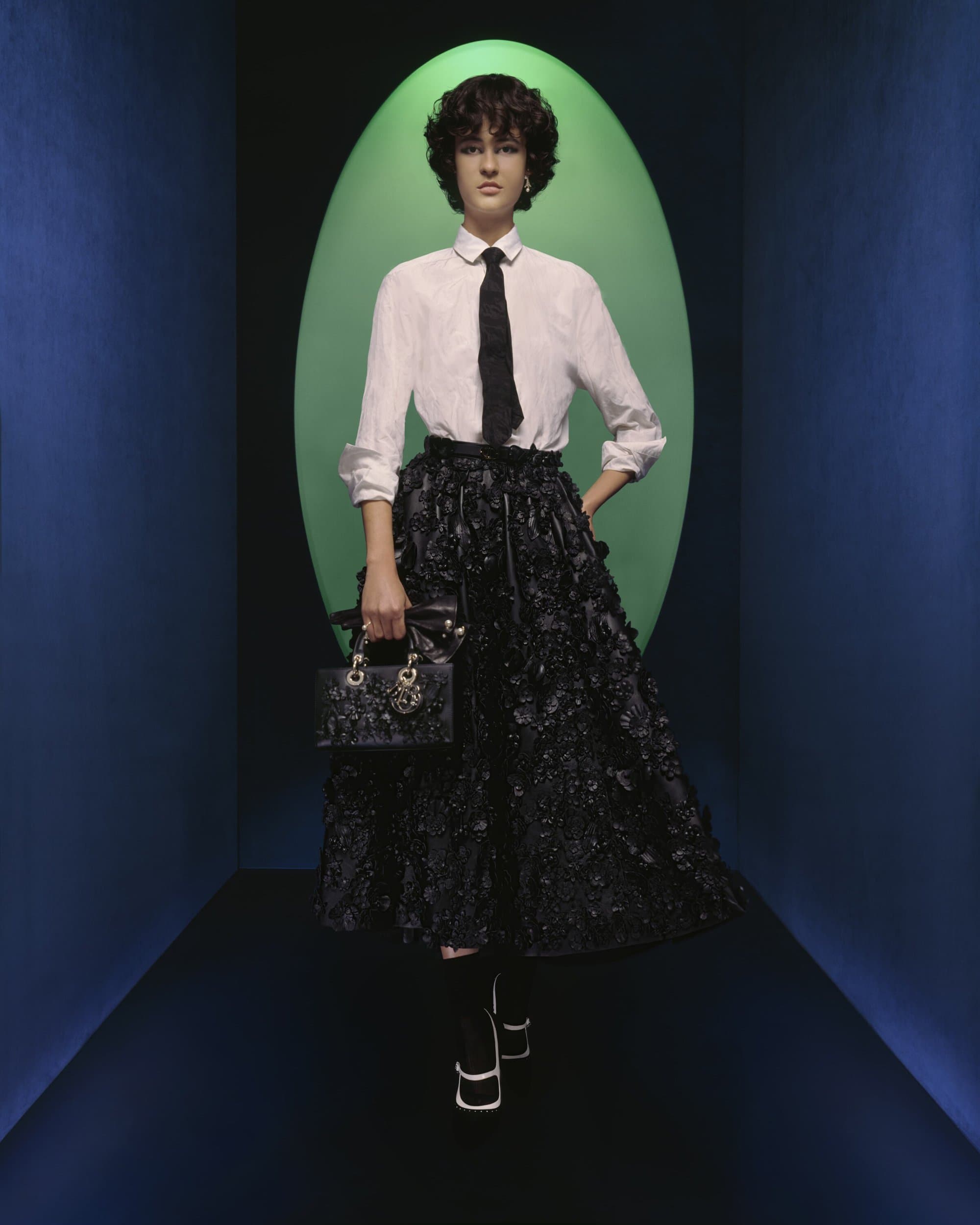 A woman stands in a fashion pose in a studio wearing a white shirt, black tie, black skirt, and carrying a black purse.