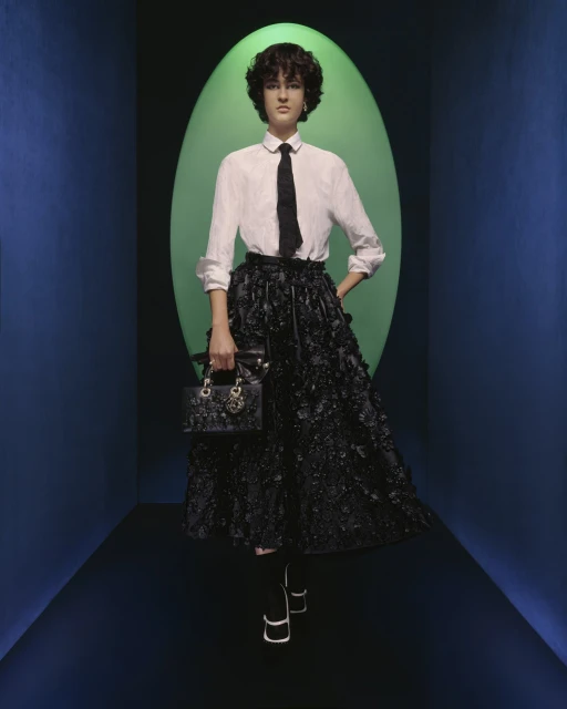 A woman stands in a fashion pose in a studio wearing a white shirt, black tie, black skirt, and carrying a black purse.