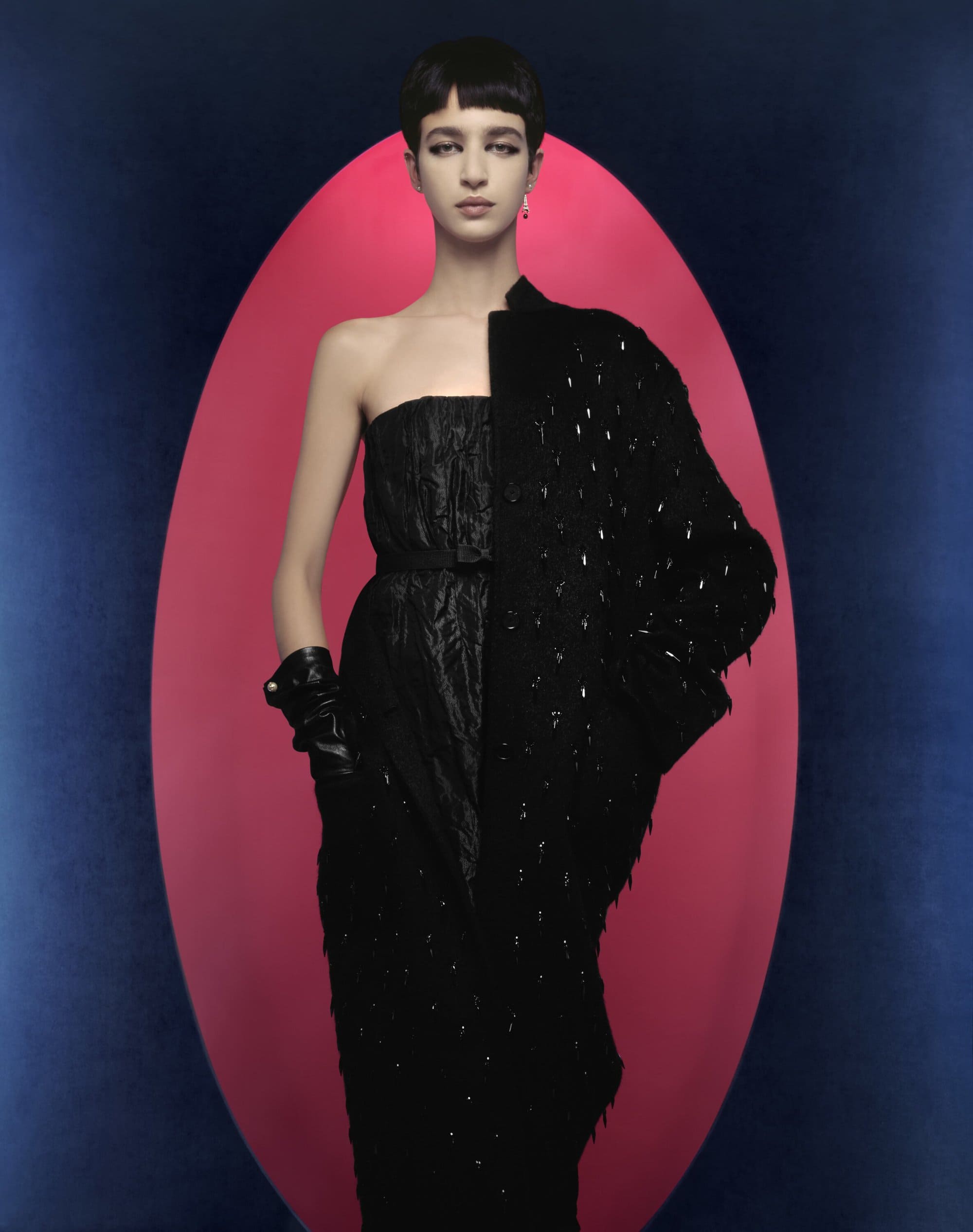 A single adult female model with short dark hair poses in a strapless black textured dress with a blue and pink oval background.