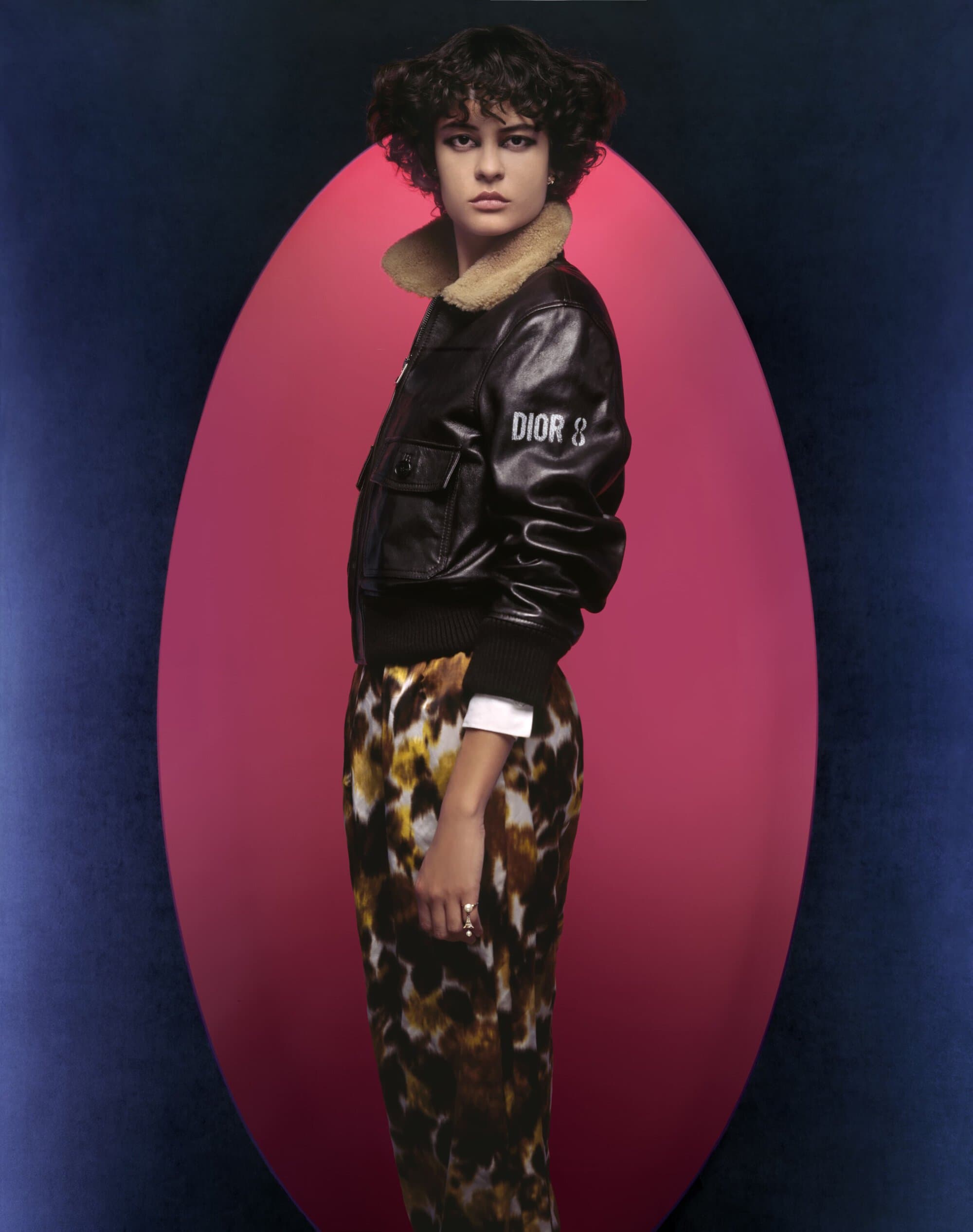 Fashionable adult female posing in a black bomber jacket with printed pants against a pink and blue backdrop in a studio setting.