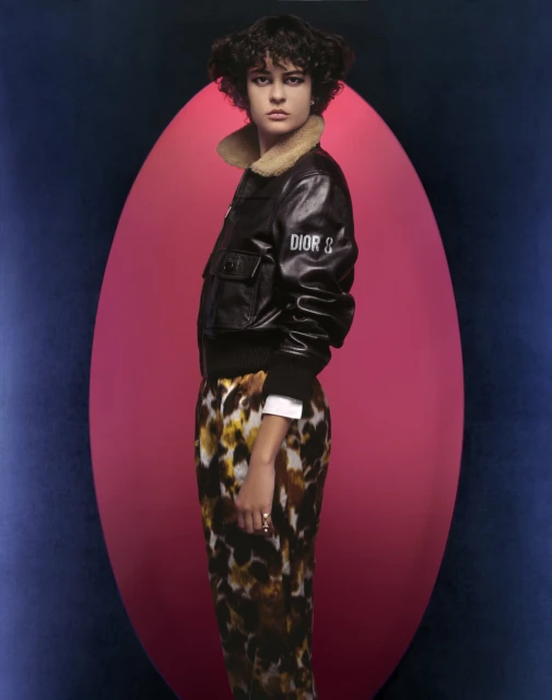 Fashionable adult female posing in a black bomber jacket with printed pants against a pink and blue backdrop in a studio setting.