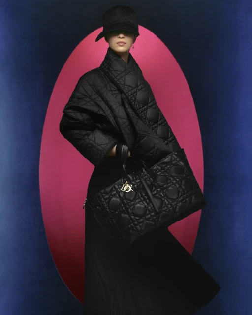 A model wearing a black quilted coat and carrying a black quilted handbag poses against a color block pink and blue backdrop.