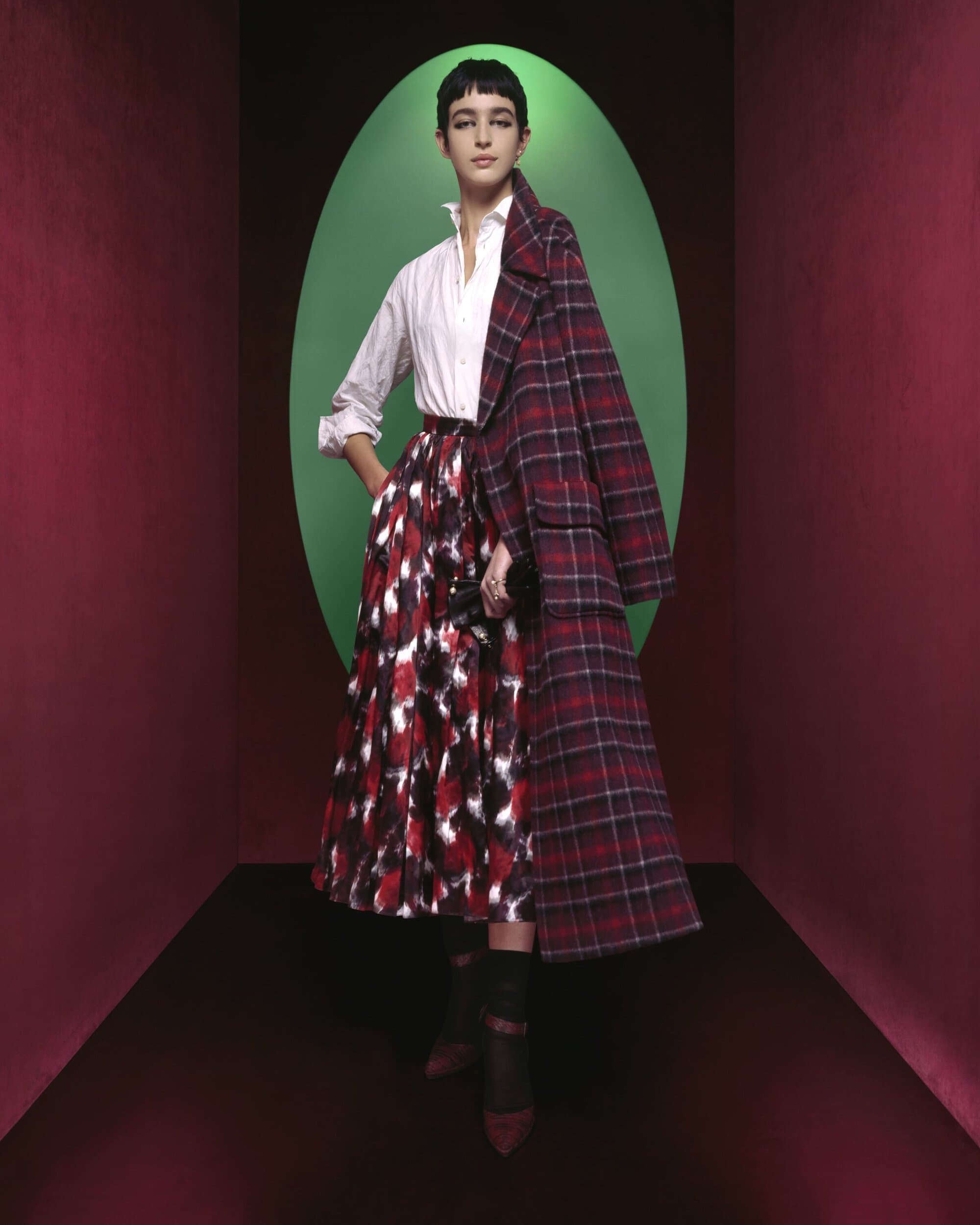 A woman in a white shirt and patterned skirt poses in front of a green oval, wearing a red plaid coat in a studio setting.